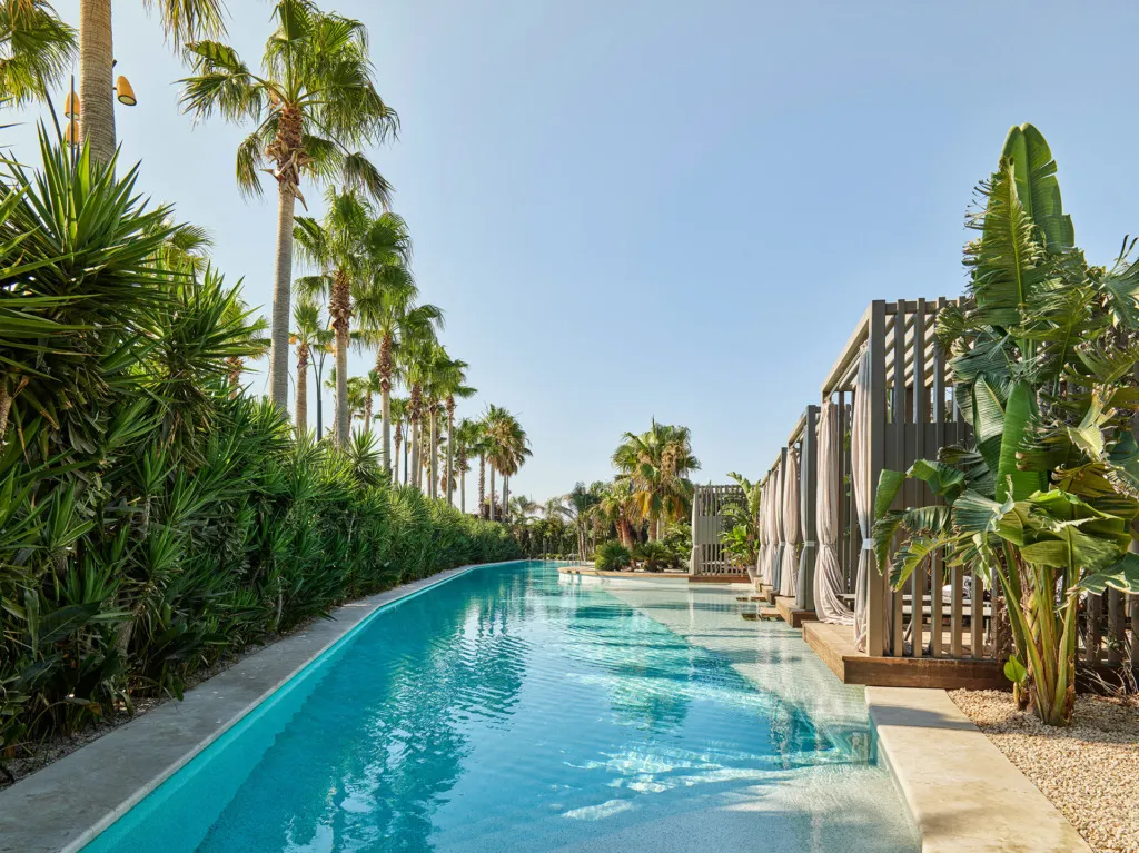 Biblos Resort Alaçatı'nın serinletici atmosferini yansıtan bu uzun yüzme havuzu, bir tarafında ahşap perdeli kabinler, diğer tarafında ise yemyeşil palmiye ağaçları ve tropik bitki örtüsüyle çevrili berrak suyuyla huzur dolu bir görüntü sergiliyor.