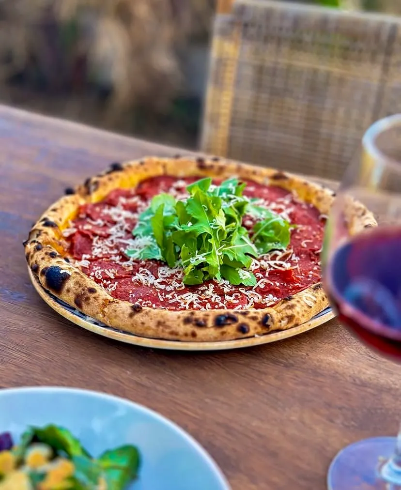 Odun ateşinde pişmiş, kenarları hafifçe yanık, bol pepperoni veya sucuk, rendelenmiş parmesan peyniri ve ortasında taze roka ile süslenmiş iştah açıcı bir pizza, ahşap masada dururken, yanında kırmızı şarap kadehi ve bulanık dış mekan manzarasıyla keyifli bir yemek anı sunuyor.
