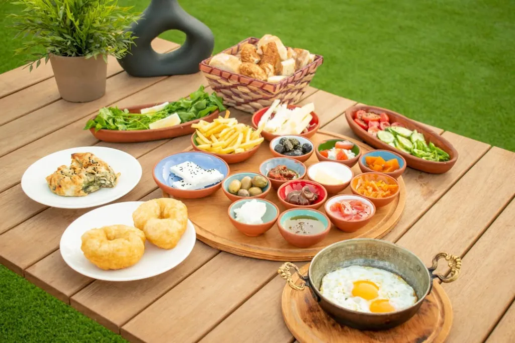 Yeşil çimler üzerinde, ahşap bir masada servis edilmiş; bakır sahanda yumurta, pişi, börek, taze ekmekler, beyaz peynir, zeytinler, tereyağı, reçeller, domates, salatalık, roka ve patates kızartması gibi birçok lezzeti barındıran, doğa içindeki zengin bir Bodrum kahvaltısı.