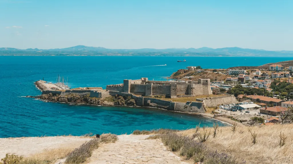 Bozcaada'nın berrak turkuaz sularıyla çevrili, tarihi kalesi ve şirin kasabasıyla birlikte uzaktan görülen bu panoramik görüntü, adanın gezilecek yerlerini keşfetmek için bir Bozcaada gezi rehberi niteliğindedir.