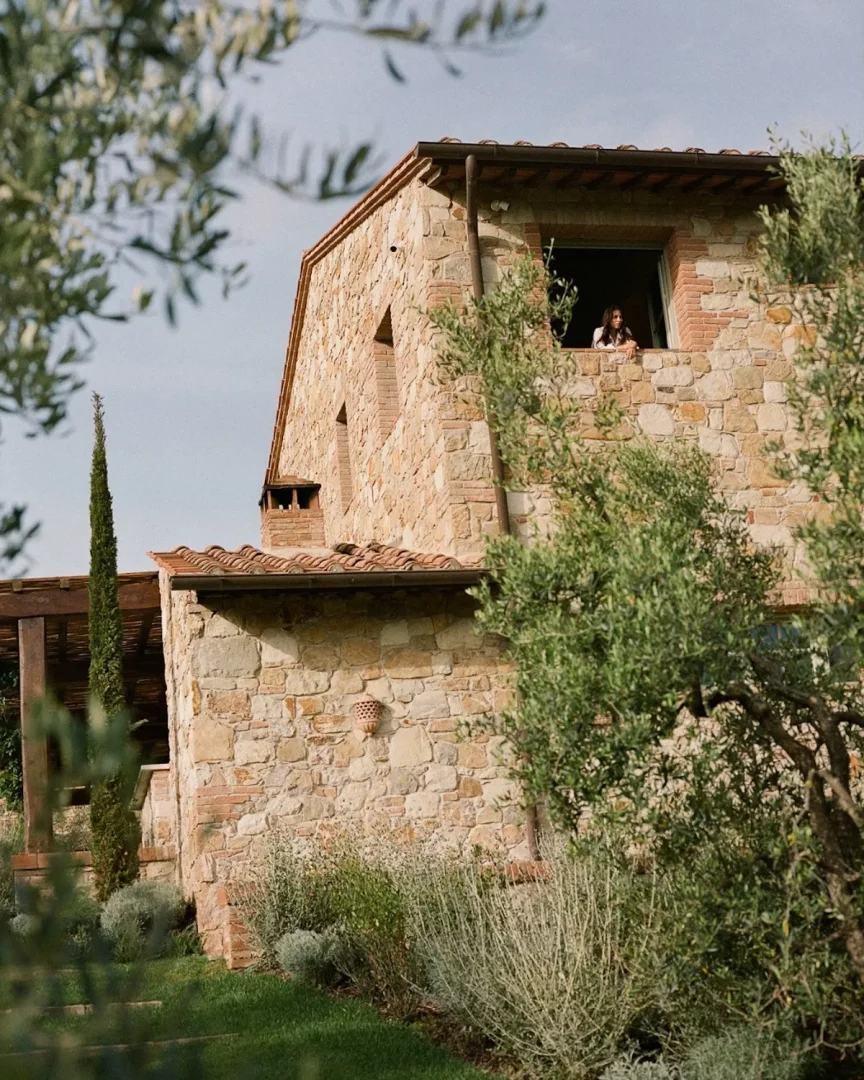 Zeytin ağaçları ve yeşil çalılıklarla çevrili, taş duvarlı ve geleneksel kiremit çatılı görkemli Castello di Casole binasının Toskana'daki bir penceresinden dışarıya doğru bakan bir kadın huzurlu manzarayı seyrediyor.