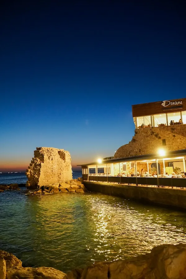 Işıkları durgun sulara yansıyan denize sıfır tarihi taş yapılı Doniana restoranında akşam yemeği yiyenler. Koyu mavi alacakaranlıkta deniz içindeki antik kalıntılar, adaların ailece yaşanacak huzurlu sahil ve lezzet kültürünü yansıtıyor.