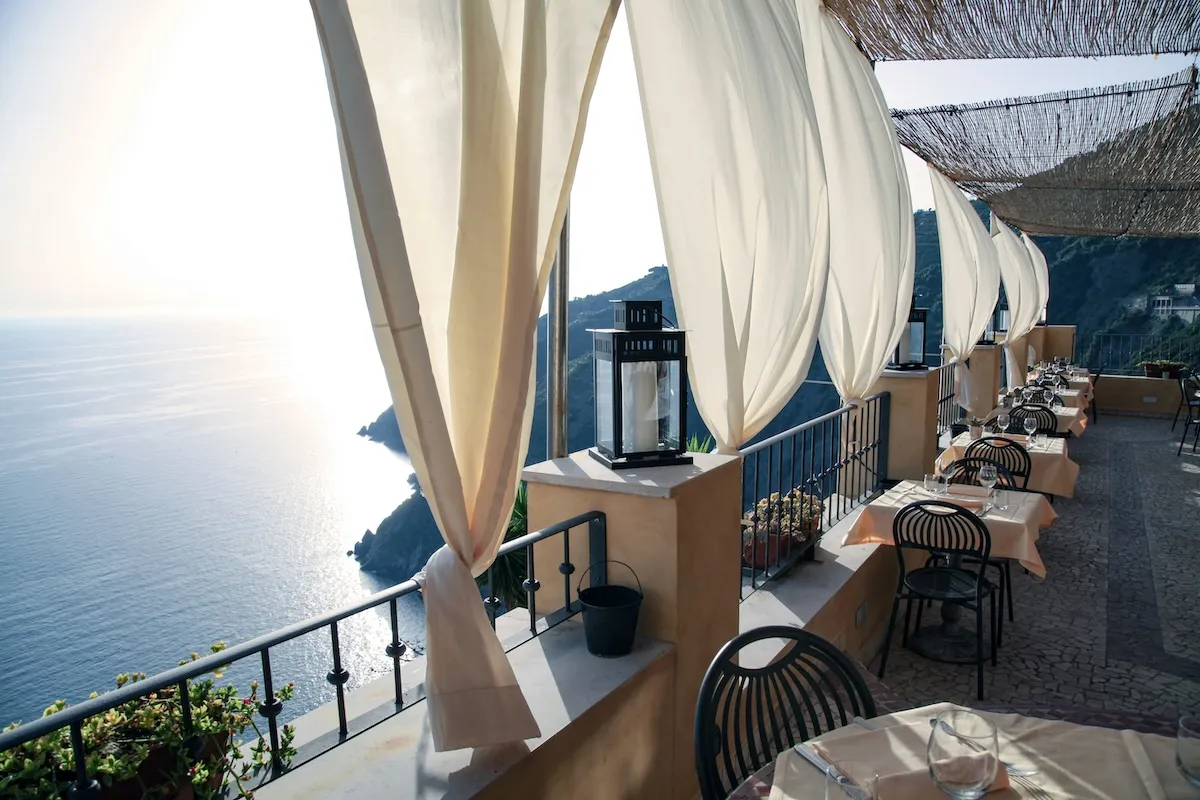 Oggusto Çocuklu Aileler İçin Yunan Adaları Rehberi: Sarp kayalıklar üzerinden parlayan denize bakan şık bir teras restoranı. Rüzgarda uçuşan beyaz perdeleri ve özenle kurulmuş masalarıyla bu mekan, adalarda ailece yenecek huzurlu ve keyifli akşam yemeklerini temsil ediyor.