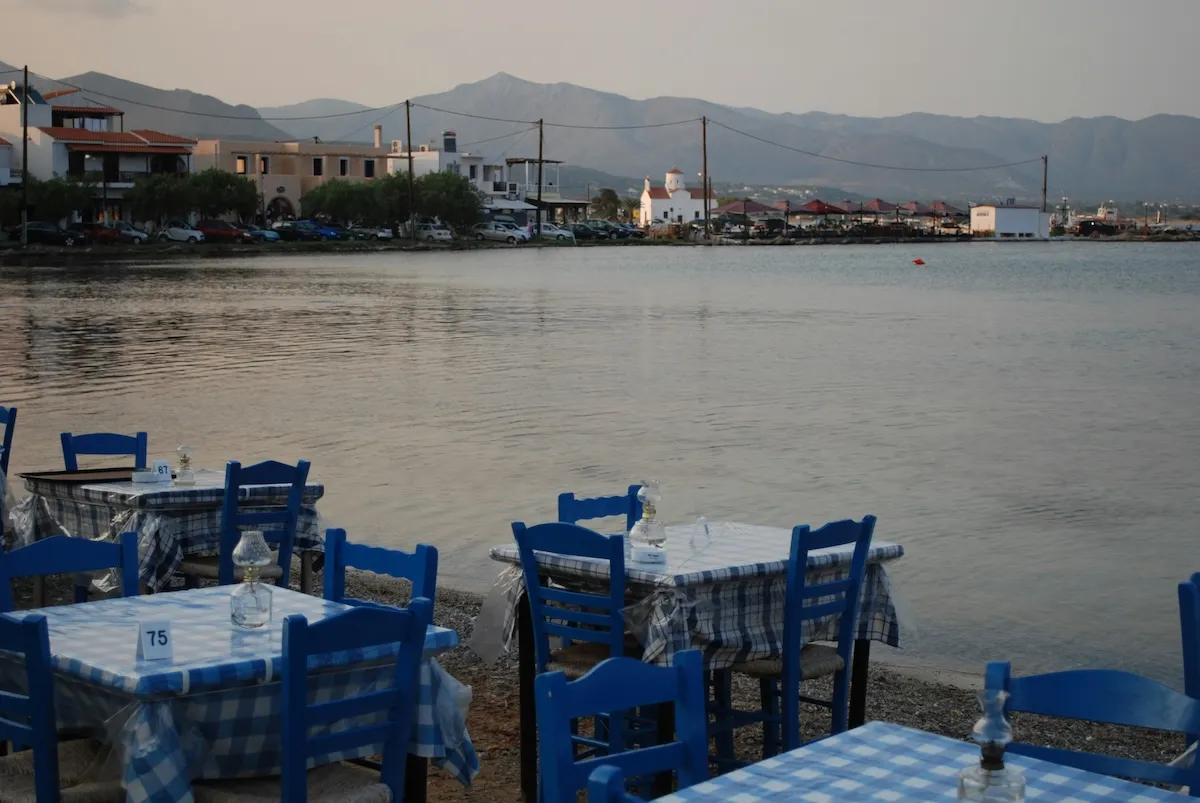 Deniz kenarında, mavi beyaz pötikareli örtüleri ve mavi ahşap sandalyeleriyle sıralanan geleneksel taverna masaları. Arka plandaki sakin koy manzarası eşliğinde, ailece yenecek huzurlu bir akşam yemeğini ve ada dinginliğini yansıtıyor.