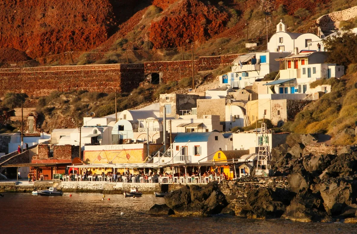 Oggusto Çocuklu Aileler İçin Yunan Adaları Rehberi: Santorini'nin dik kırmızı volkanik kayalıklarına sıralanmış beyaz badanalı geleneksel evleri. Deniz kenarındaki tavernada yemek yiyen misafirler, ailece yaşanacak huzurlu ve otantik bir ada tatilinin atmosferini yansıtıyor.