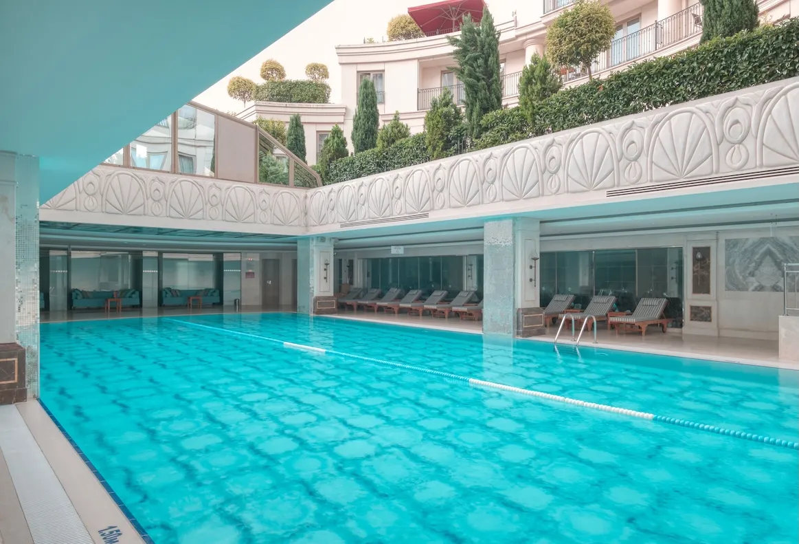CVK Park Bosphorus Hotel İstanbul'daki Safira Spa merkezine ait, parlak turkuaz renkli suyu ve şık mimarisiyle dikkat çeken, havuz kenarında sıralı şezlonglar ve cam bölmelerle ayrılmış oturma alanlarına sahip, üst kısımdaki pencerelerden dışarıdaki yeşil ağaçlar ve bina cephesinin göründüğü geniş ve aydınlık kapalı yüzme havuzu.