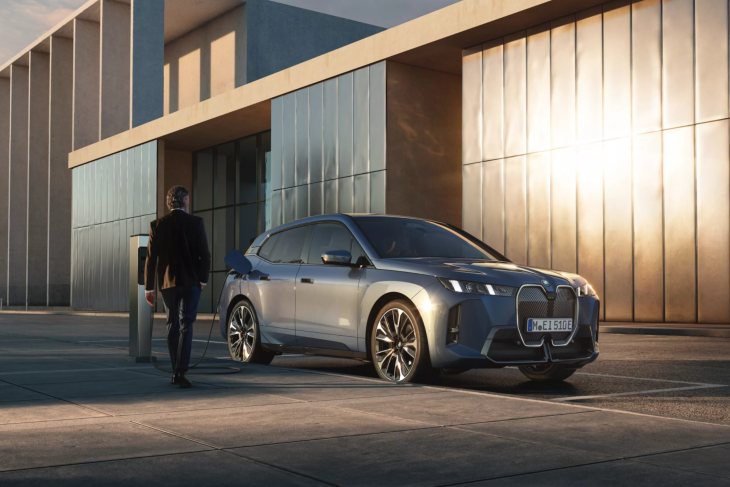 Modern mimarili bir binanın önünde, altın rengi gün batımı ışıklarıyla aydınlanmış park alanında şarj olan metalik gri bir BMW iX elektrikli SUV, şarj kablosu araca bağlıyken uzaklaşan takım elbiseli bir adamla, uzun menzilli elektrikli SUV modellerinin günlük kullanımındaki rahatlığı ve teknolojik ilerlemeyi vurguluyor.