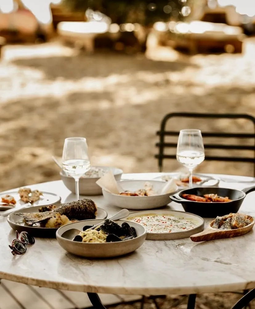 Mermer bir masada özenle dizilmiş, midyeli makarnadan ızgara balığa, çeşitli Akdeniz mezelerinden oluşan zengin bir yemek seçkisi ile iki kadeh beyaz şarabın bulunduğu, açık havada keyifli bir anı yansıtan, sıcak tonlarda çekilmiş davetkar bir görsel.