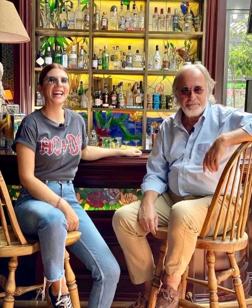Bir bar taburesinde oturan, AC/DC tişörtlü güler yüzlü bir kadın ve açık mavi gömlekli, sakallı bir adam, arkalarında renkli vitray detayları ve çeşitli şişelerle dolu bir raf ünitesi bulunan canlı bir barda keyifli bir anı paylaşıyorlar.