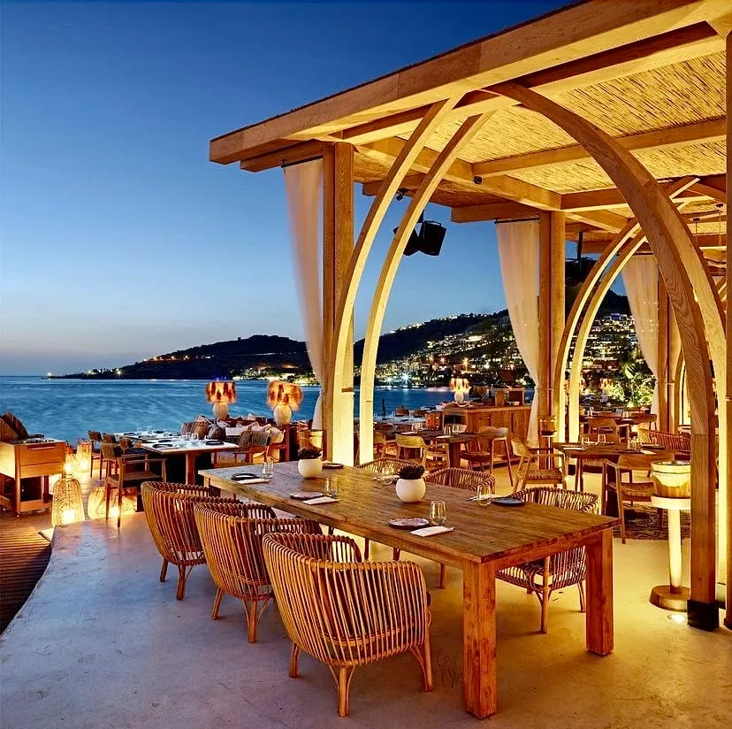 Alacakaranlıkta deniz kenarında yer alan, ahşap kemerler ve hasır çatıyla bezenmiş şık bir restoran, sıcak ışıklarla aydınlatılmış masaları, hasır sandalyeleri ve bembeyaz perdeleriyle huzurlu bir atmosfer sunarken, uzak ufukta ışıkları parlayan bir sahil kasabasının büyüleyici manzarasına hakimdir.