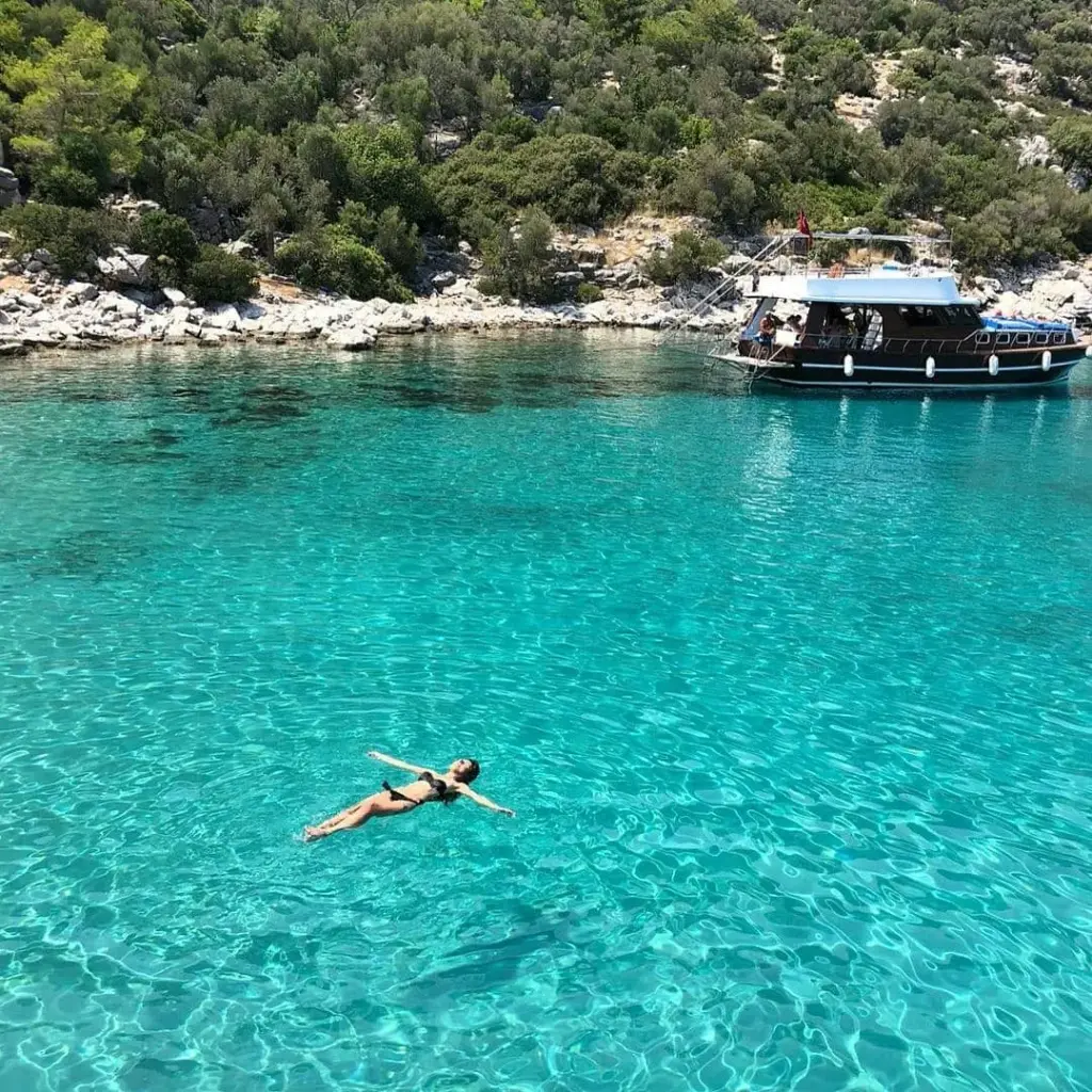 Kadın, Ege kıyılarının bakir koylarından birinde, turkuaz renkteki berrak sularda sırtüstü yüzüyor, arka planda ağaçlarla kaplı kayalık kıyıya demirlemiş bir tekne görülürken bu huzurlu ve doğal güzellik, eşsiz bir mavi yolculuk deneyimini betimliyor.
