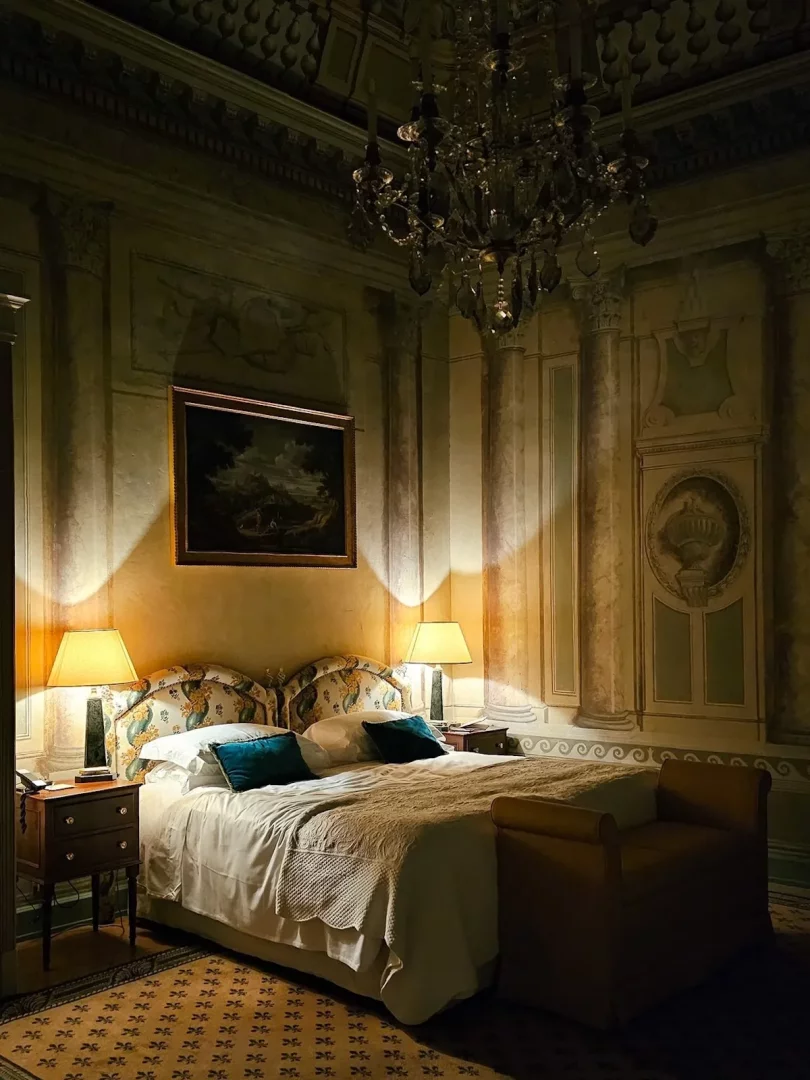 Grand Hotel Continental Siena'ya ait lüks bir otel odasında, tavandan sarkan gösterişli kristal avizenin loş ışığında, duvarlardaki soluk freskler ve sütun çizimleriyle bezeli bir atmosferde, desenli başlığı olan geniş yatak ve iki yanındaki sıcak aydınlatmalı komodin lambaları şık bir ortam sunuyor.