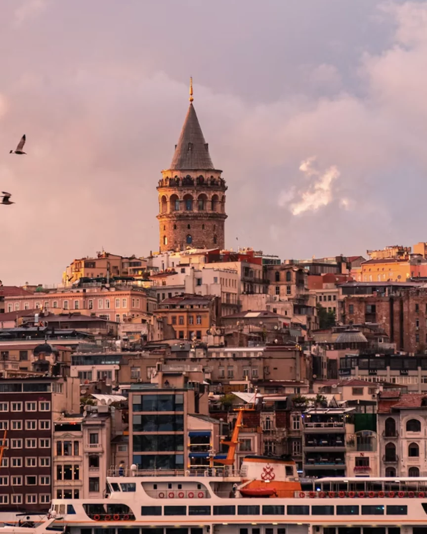 Gün batımının büyüleyici kızıl ve mor tonlarıyla aydınlanan, tarihi Galata Kulesi'nin çevresini saran kalabalık binaların arasından yükseldiği ve ön planda bir vapurun Haliç'te süzüldüğü bu İstanbul manzarası, şehrin eşsiz bir gezi rehberi deneyimi sunuyor.