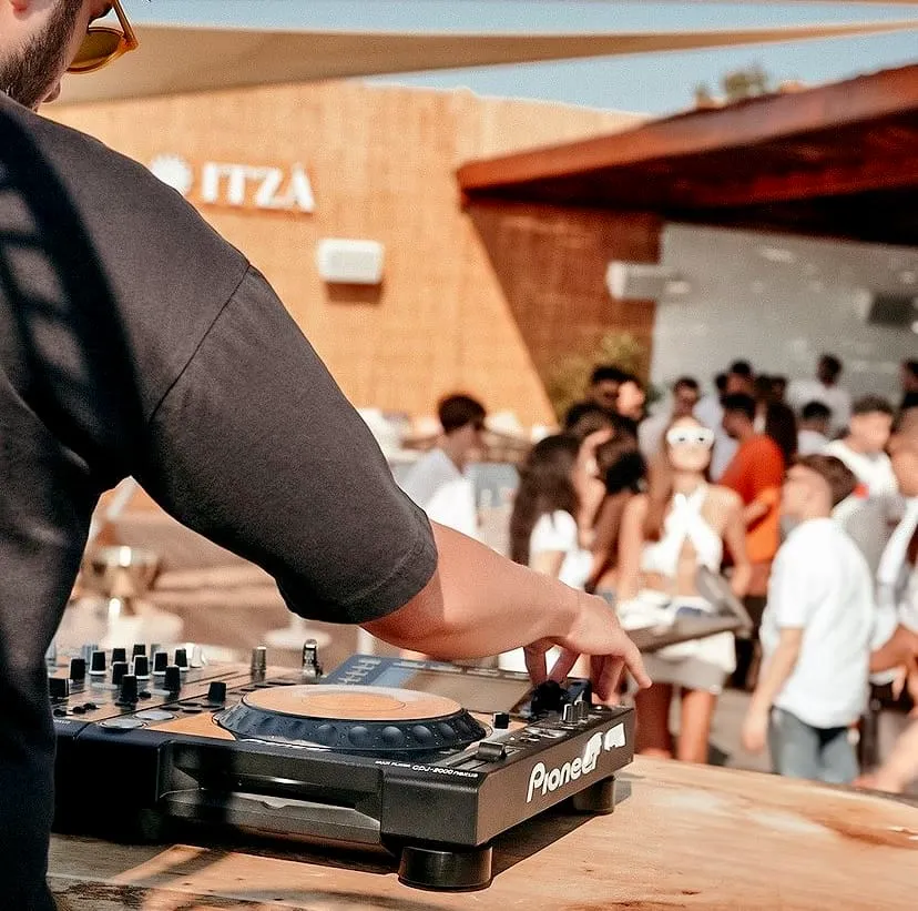 Güneşli bir ortamda, ITZA yazılı binanın önünde, güneş gözlüklü bir DJ, arkası dönük şekilde Pioneer marka profesyonel DJ ekipmanıyla müzik çalarken, arka planda canlı ve eğlenen kalabalık net bir şekilde görülüyor.