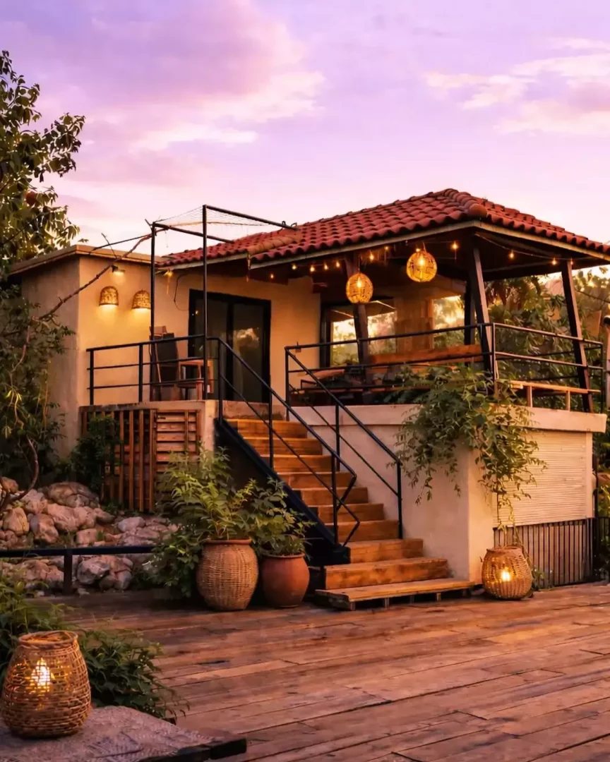 Gün batımının mor ve pembe tonlarındaki gökyüzü altında, Kaş, Kalkan, Olympos gibi bölgelerde bulunabilecek en iyi otellerden birinin görünümünde, ahşap terası, kırmızı kiremit çatısı, merdivenleri ve balkonundaki sıcak ışıklandırmalarla çevrili, dış mekan fenerleri ve bitkilerle süslenmiş davetkar bir konaklama evi.