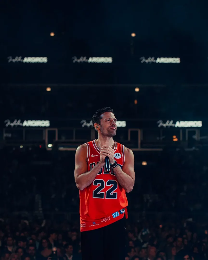 Sahnedeki Konstantinos Argiros, kırmızı renkli Bulls 22 numaralı basketbol formasını giymiş, kulağında kulaklık, mikrofonu iki eliyle göğsünün önünde tutarak yukarı bakarken, etrafını saran büyük seyirci kalabalığı ve sahne ışıklarıyla bir performans anını sergiliyor.