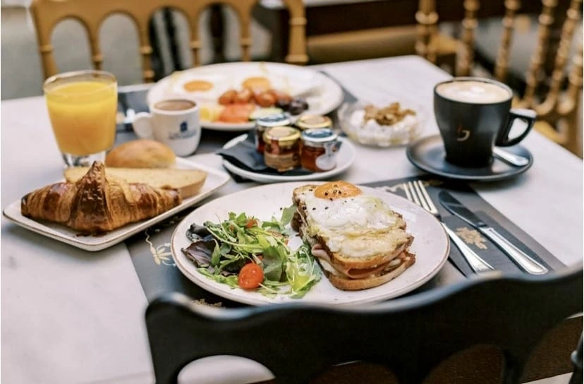 Selanik'teki Koukos'ta, sahanda yumurta ile servis edilen Croque Madame'ın yanında taze yeşil salata, kruvasan, ekmek çeşitleri, portakal suyu, kahve, farklı reçeller ve yoğurtlu kuruyemişler içeren zengin bir kahvaltı masası, zarif bir atmosferde iştah açıcı bir görüntü sunuyor.