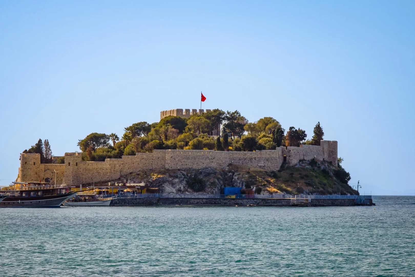 Kuşadası'nın incisi Güvercinada'daki tarihi kalenin, surlarında dalgalanan Türk bayrağı ve etrafını saran zengin yeşil bitki örtüsüyle masmavi Ege Denizi üzerinde yükselen görkemli bir görüntüsü, sol tarafta iskeleye yanaşmış tur tekneleriyle birlikte berrak bir havada resmedilmiştir.