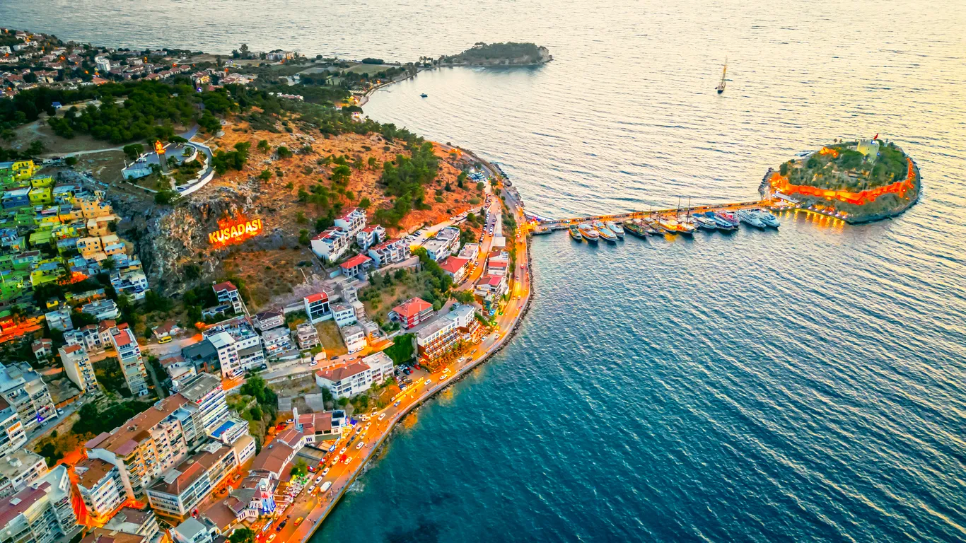 Kuşadası'nın alacakaranlıkta parlayan şehir ışıkları, kıyı şeridini saran canlı yapılar, birçok tekneye ev sahipliği yapan hareketli iskelesi ve ışıl ışıl Güvercinada'ya uzanan köprüsü, bu Ege tatil beldesine neden gidileceğine dair büyüleyici bir görsel sunuyor.