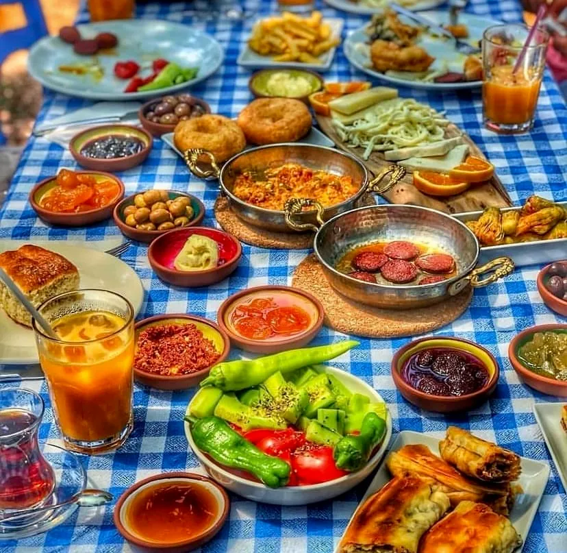 Mavi beyaz kareli bir masa örtüsünde, menemen, sucuk, çeşitli peynirler, zeytinler, reçeller, kızarmış hamur işleri, çıtır börekler, taze sebzeler ve portakal suyu ile çay gibi birçok lezzeti bir araya getiren zengin ve geleneksel bir Türk kahvaltısı sofrası, kuytu bir mekanın sıcak ve otantik atmosferini başarıyla vurguluyor.