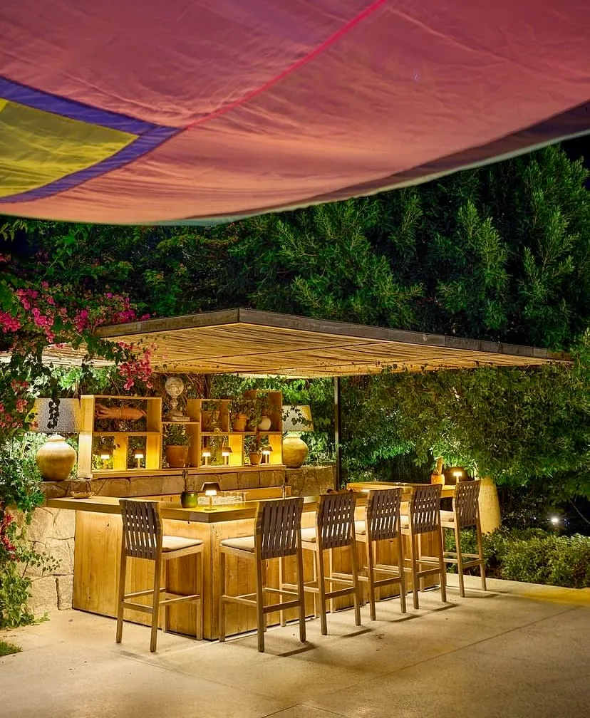 Gece çekilmiş bu dış mekan görüntüsünde, sıcak ışıklarla aydınlatılmış şık ahşap bir bar ve üzerinde minderli beş adet ahşap bar taburesi, arkasında bitkiler, dekoratif objeler ve aydınlatmalarla dolu raflar yer alıyor; üstte renkli bir tente ve etrafta yemyeşil ağaçlar ile pembe çiçekli sarmaşıklar, Maça Kızı'nın karakteristik lüks ve doğal atmosferini yansıtıyor.