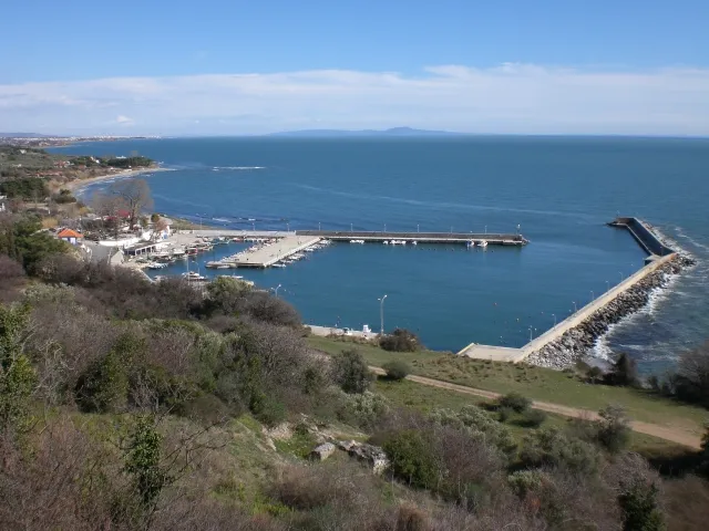 Evros bölgesindeki Makri Limanı'nın ve kıyı şeridinin kuşbakışı görünümü, mendireklerle korunan liman içinde demirli çok sayıda tekneyi, kavisli kumsalı ve arka planda masmavi denizi gösterirken, ön planda seyrek bitki örtülü ve ağaçlık bir yamaç yer alıyor.
