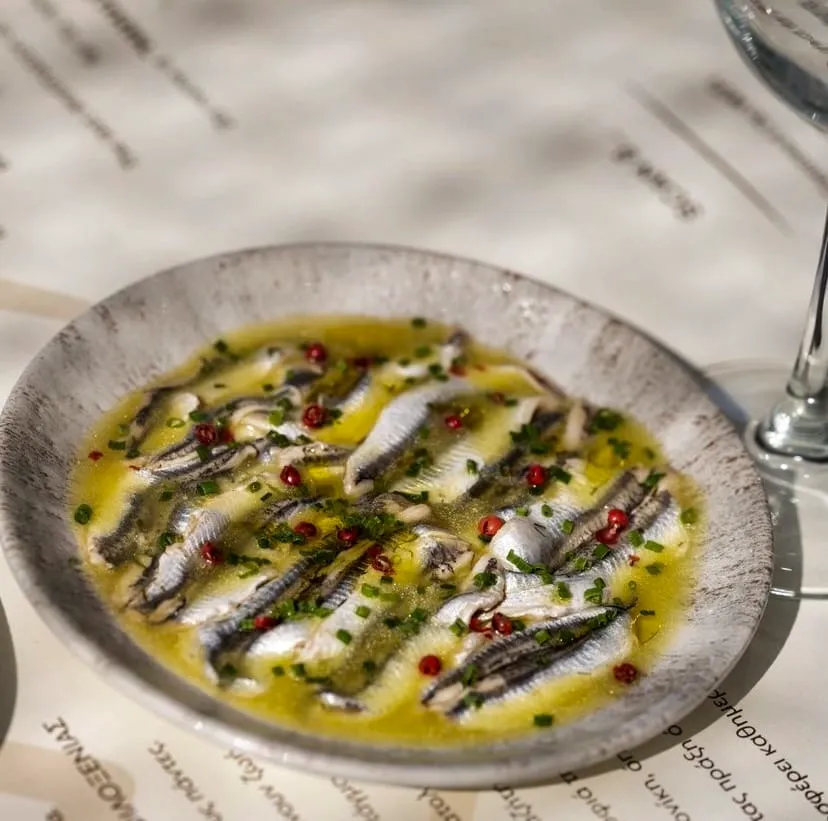 Gri ve dokulu oval bir tabakta, Selanik'in meşhur Mamalouka restoranında sunulan, zeytinyağlı marinasyon içinde bekletilmiş, üstüne ince doğranmış taze soğan ve pembe tane karabiber serpilmiş parlak gümüş renkli marine ançuezler, yazıların okunabildiği beyaz bir masa üzerinde iştah açıcı bir meze olarak yer alıyor.