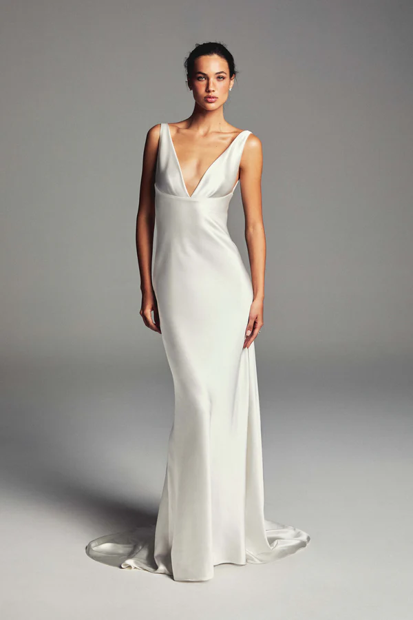 Monique Lhuillier’in modern ve zamansız tasarım anlayışını yansıtan, derin V yakalı, parlak saten kumaştan yapılmış, vücudu kusursuzca saran ve yere doğru akıcı bir şekilde uzanan minimalist bir gelinlik modeli, sade fon önünde zarif bir duruş sergiliyor.