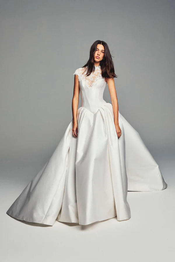 Monique Lhuillier tasarımı bu ihtişamlı gelinlik, dantel işlemeli yüksek yakası, kap kol detayları ve heykelsi bir duruş sergileyen hacimli, kıvrımlı saten eteğiyle modern zarafeti ve couture dokunuşlarını bir araya getiriyor.