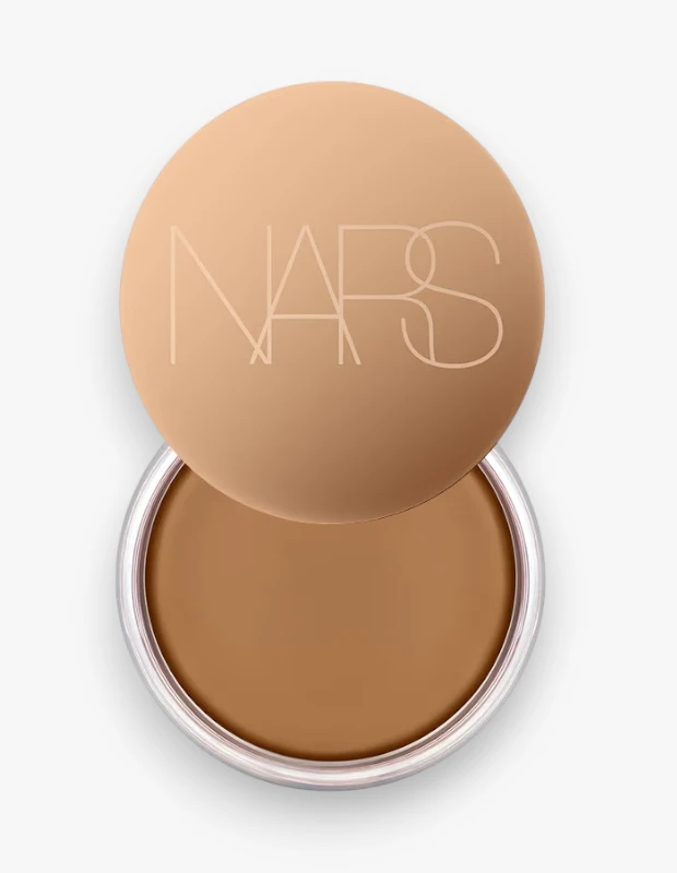 Açık bej renkli kapağında NARS logosu olan, kapağı hafifçe açık duran ve içinde pürüzsüz, yoğun kahverengi tonda NARS Laguna Bronzlaştırıcı Krem'i gösteren bir ürün kutusunun yakından görünümü.