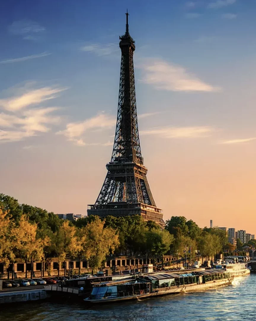 Eyfel Kulesi'nin altın rengi gün batımı ışıkları altında Seine Nehri kıyısındaki ağaçlar ve gezinti tekneleriyle göründüğü Paris manzarası, Babalar Günü tatil önerileri için sanat ve şehir rotası ilhamı sunuyor.