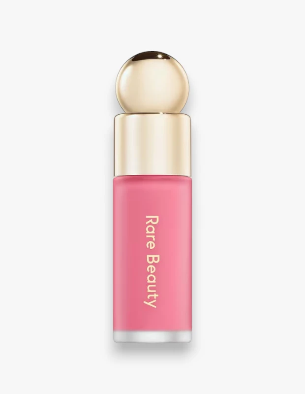 Rare Beauty Soft Pinch mini likit allık, parlak altın rengi küresel kapağı ve Rare Beauty markasının dikey olarak yazılı olduğu pastel pembe tonlarındaki mat şişesiyle beyaz bir zeminde dikkat çekici bir şekilde sergileniyor.