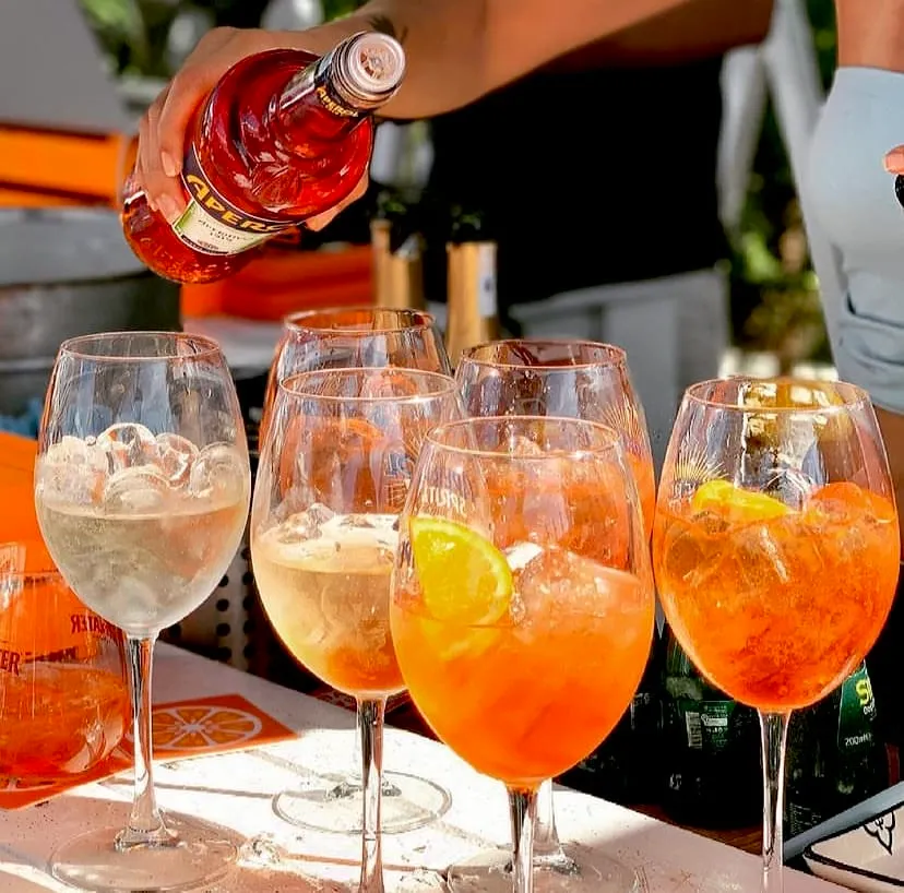 Güneşli bir havada, bir el Aperol şişesinden buz ve dilimlenmiş narenciye ile dolu şarap kadehine parlak turuncu Aperol döküyor; masada ise farklı renklerdeki diğer serinletici Aperol Spritz kokteylleri yazın tadını çıkarmak için bekliyor.