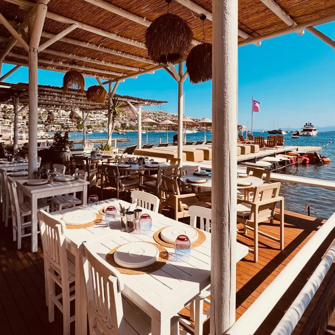 Güneşli bir Akdeniz gününde, mavi denizin ve koydaki teknelerin yanında, hasır çatılı ve rustik lambalarla süslü beyaz ahşap iskele üzerindeki şık açık hava restoranında, yemek için özenle hazırlanmış masalar.