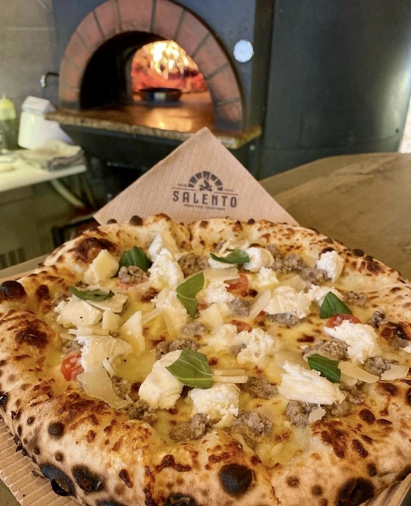 Selanik'teki Salento'dan servis edilmiş, kenarları hafifçe kömürleşmiş, üzerinde bol sosis, kremsi ricotta, rendelenmiş parmesan peyniri, domates parçacıkları ve taze fesleğen yaprakları bulunan, arka planda alevli taş fırının görüldüğü ağız sulandırıcı bir pizza.