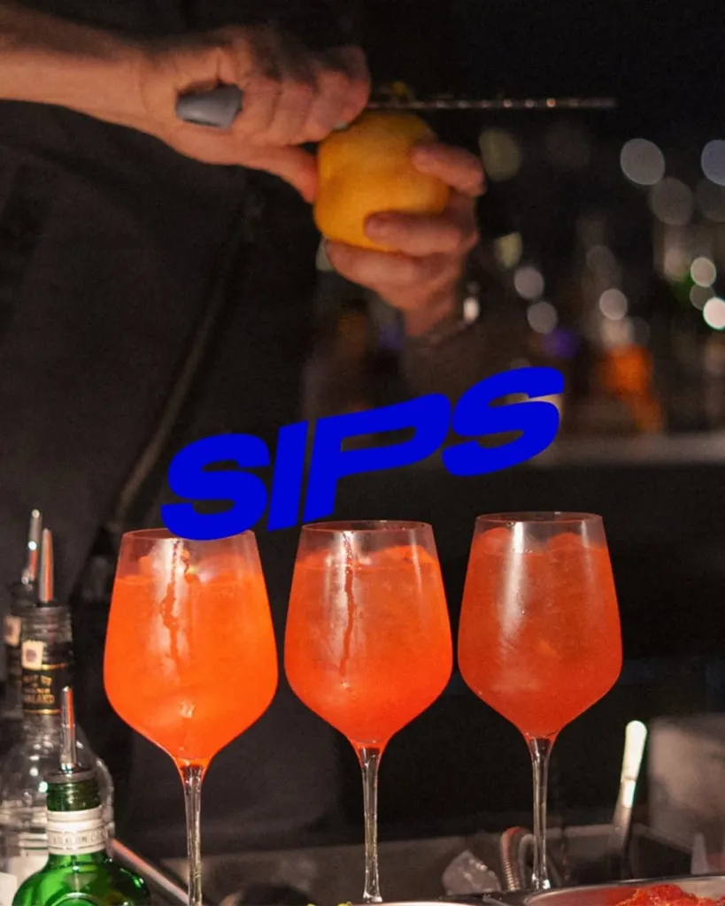 Neon mavi SIPS yazısıyla ön plana çıkan barmen, koyu renkli bir barda taze limon kabuğunu rendeleyerek, buzlu ve parlak turuncu renkteki üç zarif kokteyl kadehine son dokunuşu yaparken görülüyor.