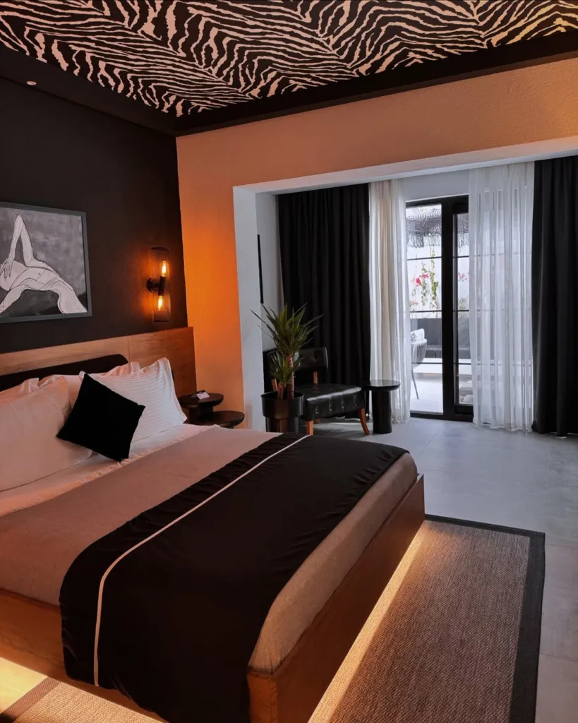 Zebra desenli tavanıyla dikkat çeken bu şık otel odasında, ahşap baza altından yayılan sıcak ışıklandırmanın aydınlattığı geniş yatak, koyu renk mobilyalar ve dışarıya açılan pencere modern bir konaklama deneyimi sunuyor.