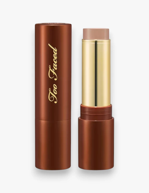 Too Faced markasına ait, çikolata kahverengisi silindirik ambalajlı ve altın detaylı, açık bej renginde bir krem stick makyaj ürünü; kapalı ve kapağı açık haliyle beyaz zeminde dikkat çekici bir şekilde sergileniyor.