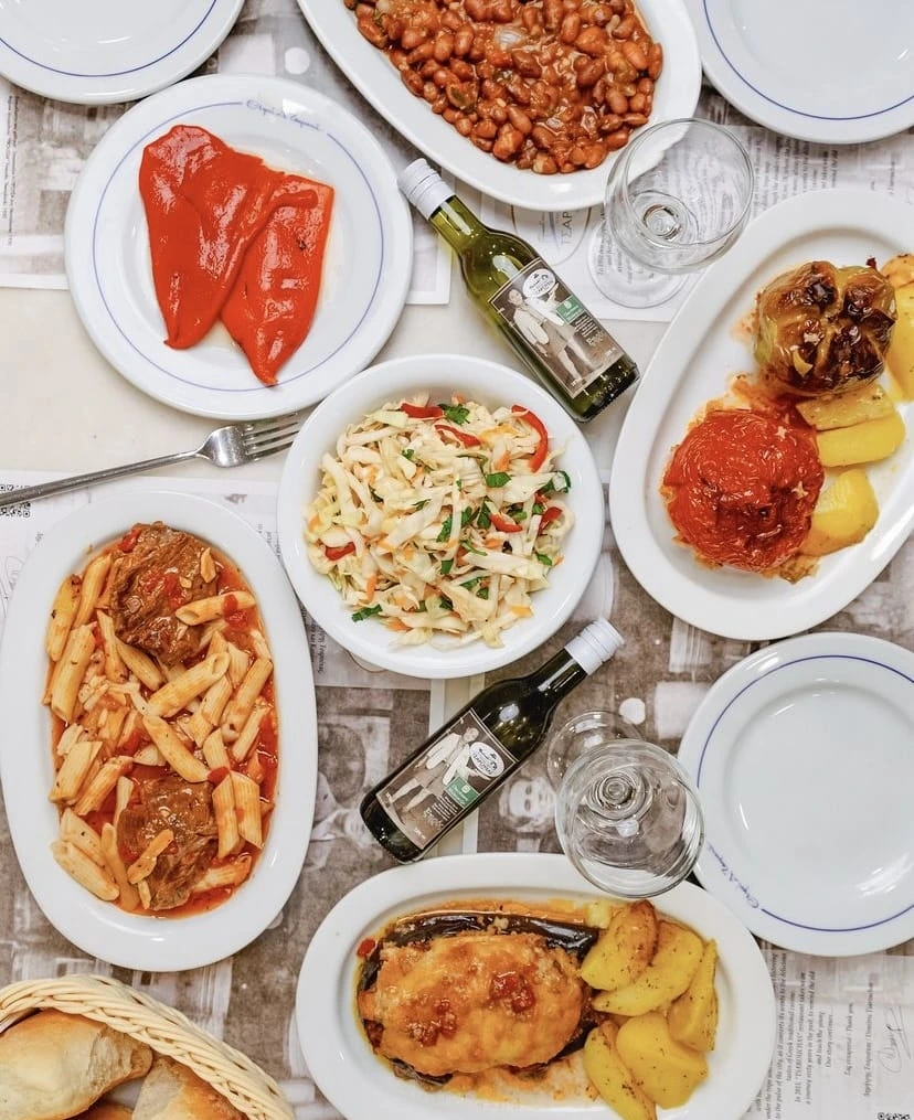 Selanik'teki geleneksel bir tavernada sunulan; kavrulmuş kırmızı biberler, kuru fasulye pilaki, lahana salatası, etli penne makarna, domates ve biber dolmaları ile patatesli patlıcan yemeği gibi çeşitli otantik Yunan lezzetleri ve iki şişe beyaz şarapla dolu, tepeden çekilmiş zengin bir masa görünümü.