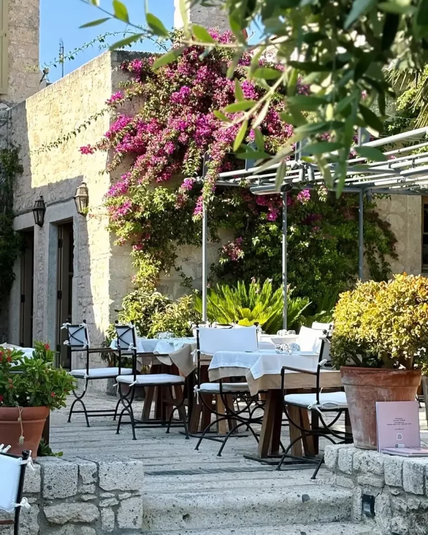 Taş bir binanın pembe begonvillerle süslü duvarının önünde, beyaz örtülü masalar ve sade sandalyelerle düzenlenmiş, dış mekanda keyifli bir yemek deneyimi sunan, seyahat etmeye değer bir restoranın Alaçatı atmosferini yansıtan davetkar terası.