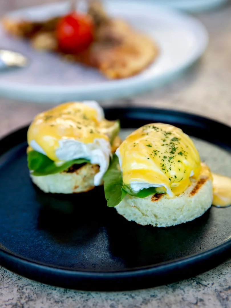 Siyah bir tabakta, kızarmış muffinler üzerinde taze yeşillikler, haşlanmış akışkan yumurtalar ve üzerine otlar serpilmiş parlak sarı hollandaise sosuyla hazırlanmış iki adet Eggs Benedict yemeği net bir şekilde görünüyor; arka planda ise bulanık bir şekilde başka bir yemek tabağı seçilebiliyor.