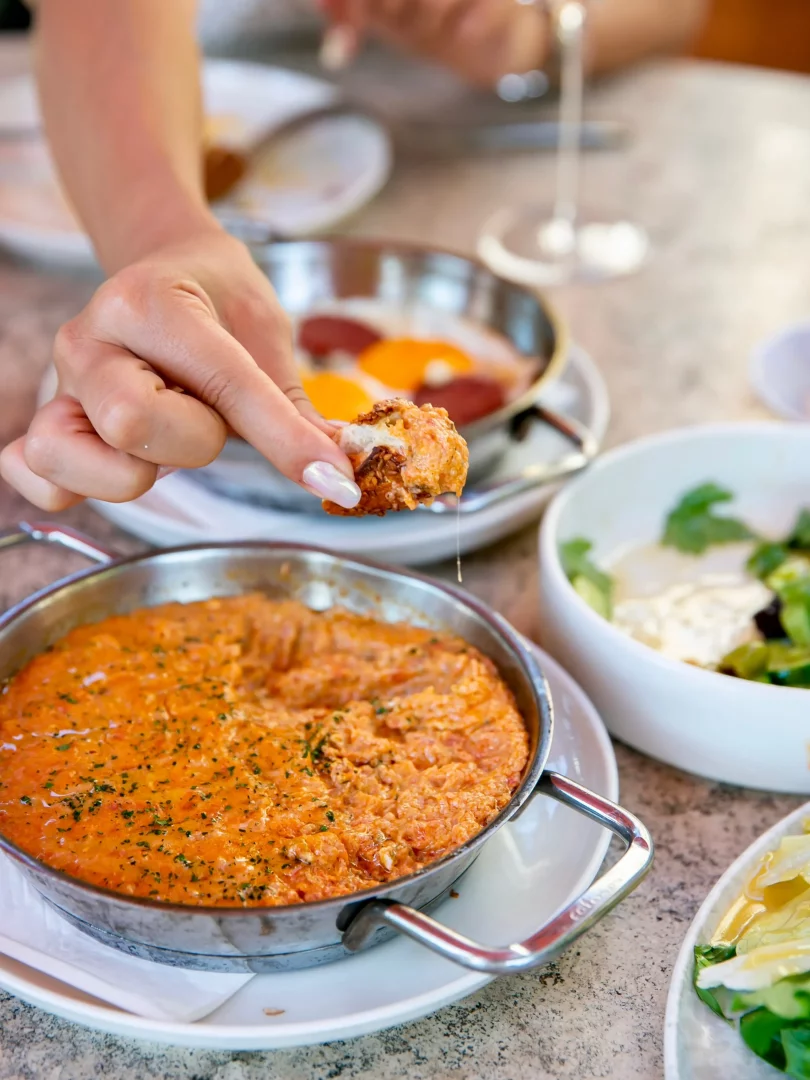 Bir kişinin parlak oje sürmüş eli, kahvaltı masasında, ön plandaki demir sahanda nefis görünen domatesli ve yumurtalı menemen yemeğinden, arkadaki sucuklu yumurta ve salataların eşlik ettiği zengin bir Türk kahvaltısı sırasında lokma alırken damlayan sosla birlikte gösteriliyor.