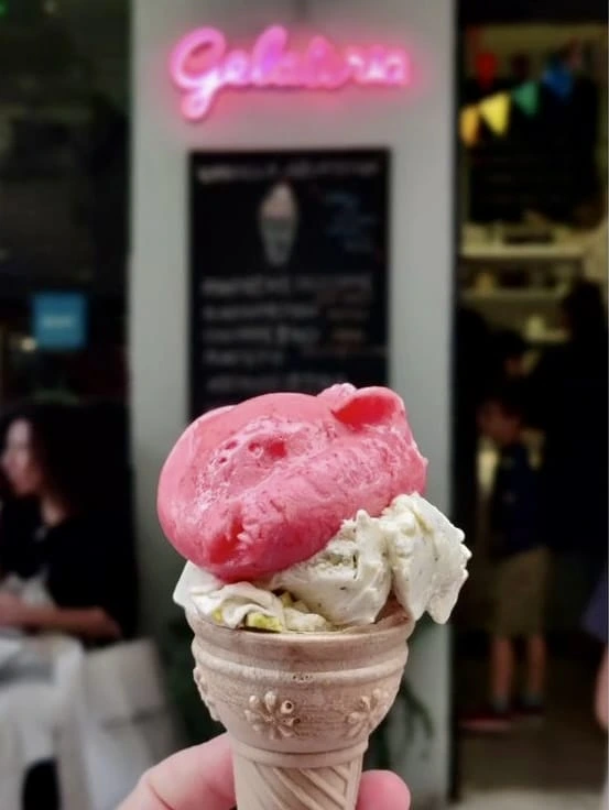 Elinde, üstünde pembe bir top ve altında vanilya veya fıstık dondurması olan, desenli bir külahı tutan kişinin yakın çekim fotoğrafında, arka planda pembe neon Gelateria tabelası ve bulanık bir dondurmacı dükkanı görülüyor.