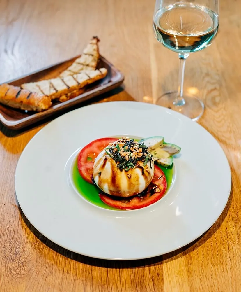 Beyaz ahşap bir masa üzerinde, üzerine koyu sos gezdirilmiş, fesleğen ve fındık parçacıklarıyla süslenmiş yuvarlak bir burrata peynirinin, dilimlenmiş domates ve canlı yeşil sos eşliğinde beyaz tabakta iştah açıcı bir şekilde sunulduğu, arka planda ızgara ekmek dilimleri ve beyaz şarap kadehiyle tamamlanan bu göz alıcı özel lezzet, damaklarda unutulmaz bir iz bırakmaya hazır.