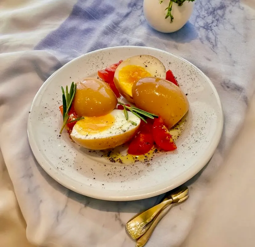 Beyaz bir tabakta, geleneksel Selanik yumurtası veya zeme yumurtası olarak bilinen, dışı kararmış ve içi kremsi sarı renkte pişmiş haşlanmış yumurtalar özenle yerleştirilmiş. Yumurtaların bazıları ikiye bölünmüş olup, hafif akışkan parlak sarı yumurta sarısı belirgin bir şekilde görülüyor. Tabağın etrafında taze doğranmış kırmızı domatesler, aromatik taze biberiye dalları, zeytinyağı ve karabiber serpilmiş; bu iştah açıcı sunum kahvaltı sofraları için ideal.