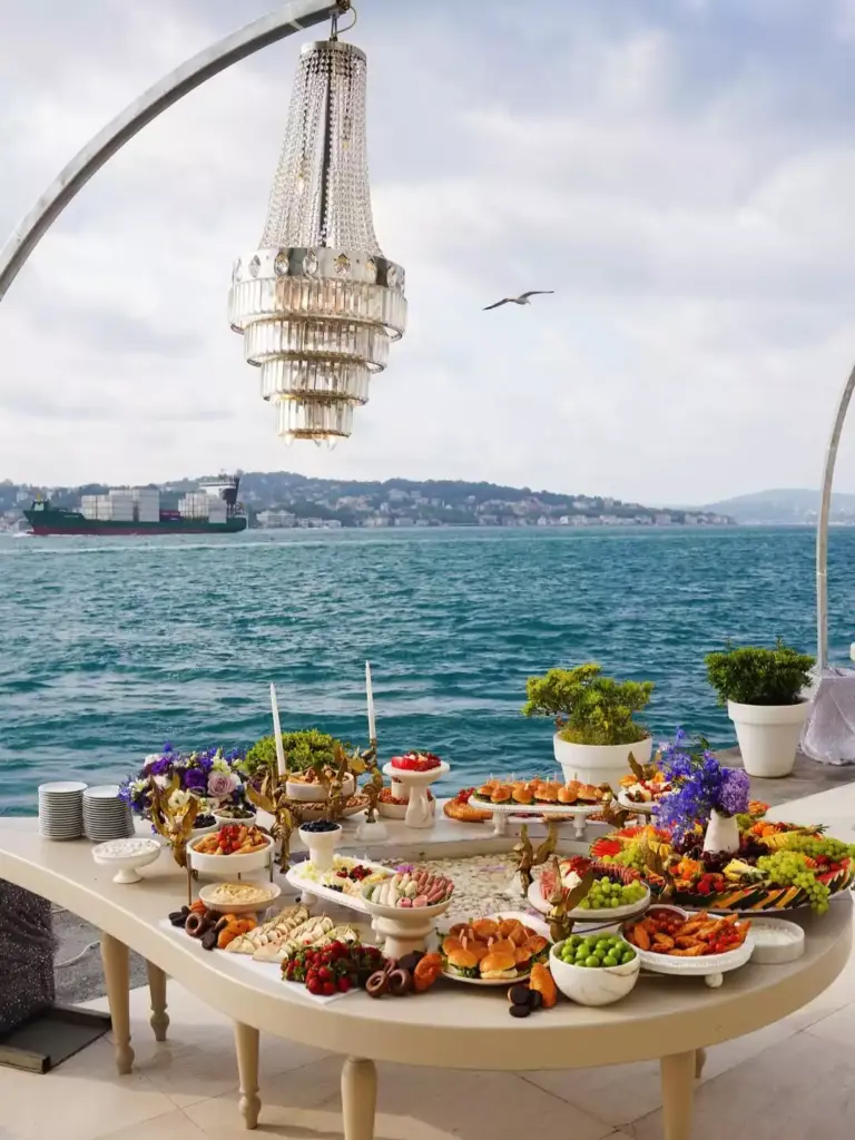 Ajia Hotel'in Boğaz manzaralı terasında, üzerinde şık kanepeler, meyveler ve çeşitli ikramlarla donatılmış uzun bir ziyafet masası, havada asılı duran kristal avize ve uzakta geçen gemilerle büyüleyici bir davet ortamı sergileniyor.