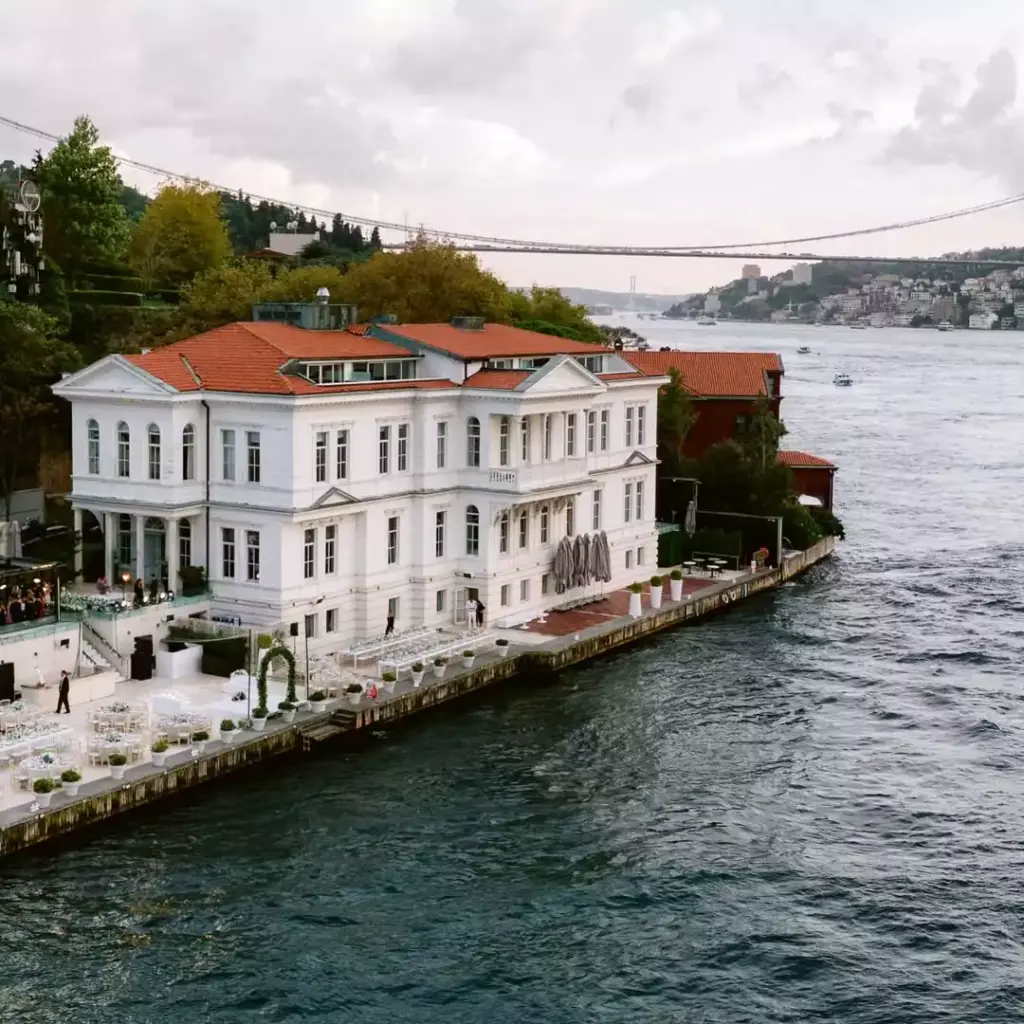 Boğaz'ın kıyısında konumlanmış, kırmızı kiremit çatılı, beyaz ve görkemli Ajia Hotel, su üzerindeki teraslarında şık açık hava etkinlik ve yemek düzenlemeleriyle dikkat çekerken, arka planda Boğaz Köprüsü ve karşı yaka manzarası uzanıyor.