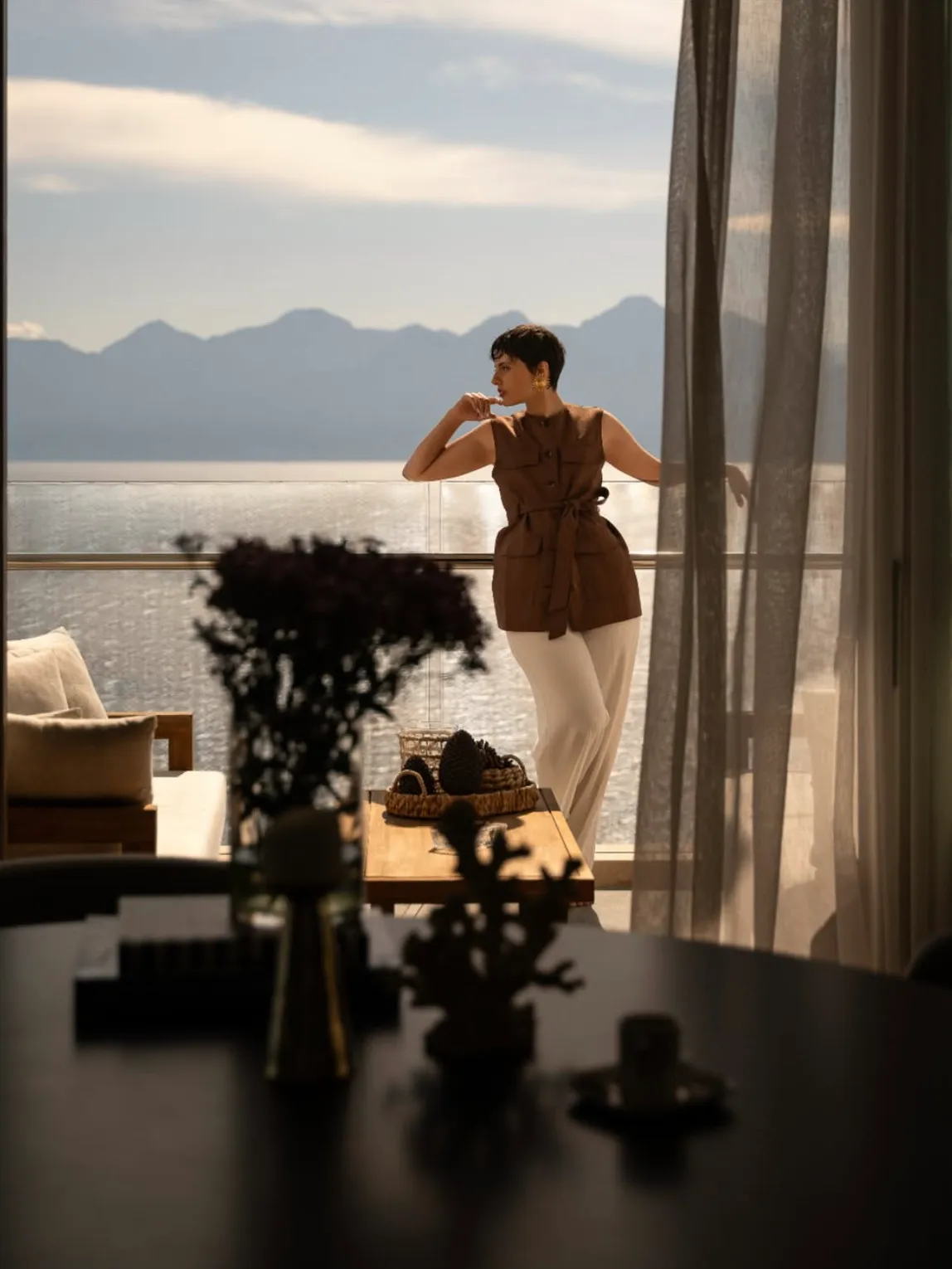 Kısa saçlı, kahverengi kolsuz bluz ve açık renk pantolon giyen bir kadın, Akra Hotel'in şık balkonunda cam korkuluğa yaslanmış, düşünceli bir ifadeyle engin denizi ve ufuktaki görkemli dağ sıralarını seyrediyor.