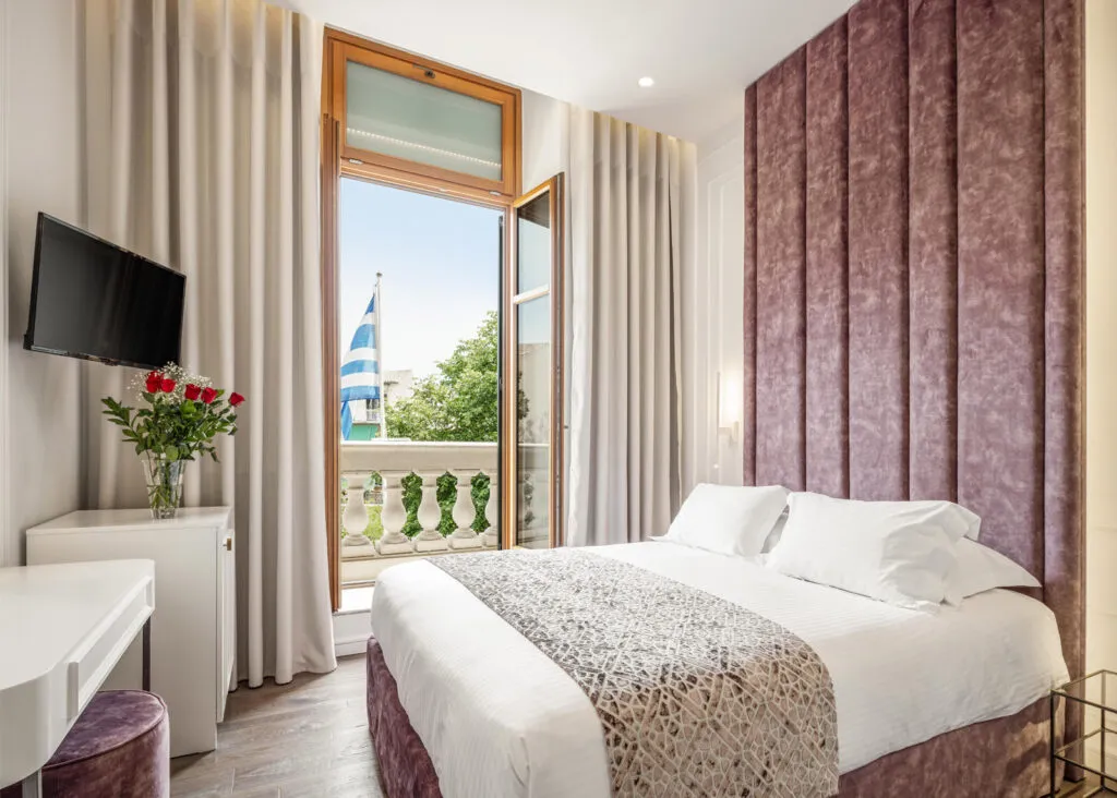 Şık döşenmiş otel odasında, mor kadife panelli yatak başlığına sahip beyaz nevresimli çift kişilik yatak ve açık balkon kapısından görünen, üzerinde Yunan bayrağının dalgalandığı, yeşillik ve masmavi gökyüzü manzarası huzurlu bir atmosfer yaratıyor; odanın solunda ise duvara asılı TV ve kırmızı güllerle dolu şeffaf vazo estetik bir detay sunuyor.
