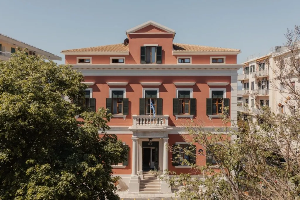 Beyaz sütunlu görkemli girişi, balkonundan sarkan küçük Yunan bayrağı ve yeşil panjurlu pencereleriyle Bella Venezia oteli veya tarihi binası, kırmızımsı turuncu rengi ve çevresindeki yemyeşil ağaçlar eşliğinde berrak mavi gökyüzü altında zarifçe yükseliyor.